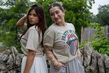 Two women standing outdoors with a stone wall and greenery in the background