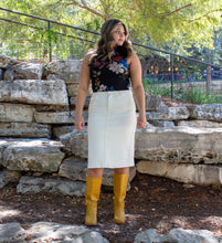 Woman standing in front of a stone wall wearing a floral top, white skirt, and yellow rain boots.